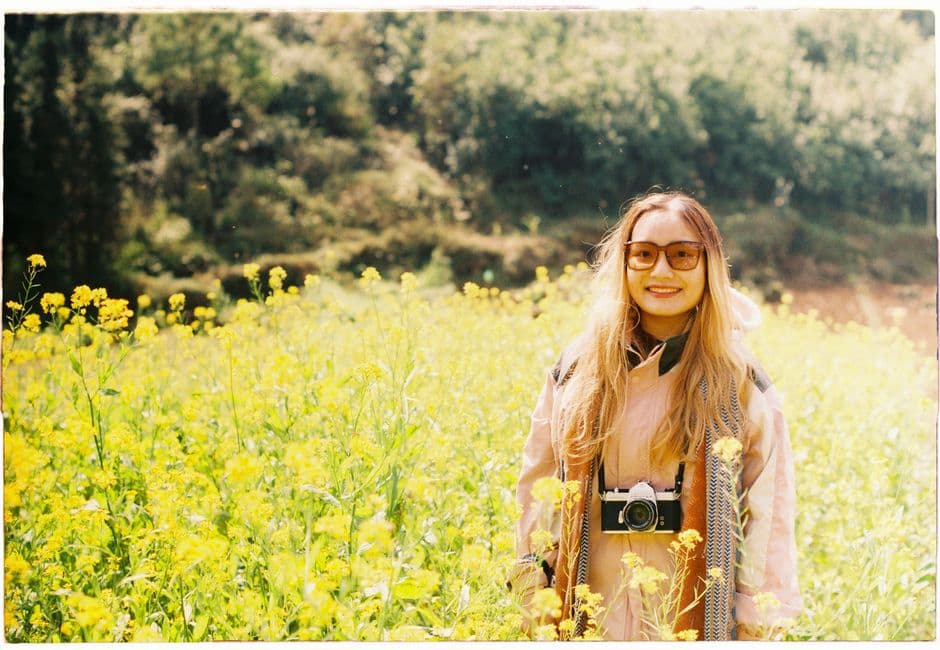 Smiling woman with camera standing in a sunlit field of wildflowers wearing sunglasses.