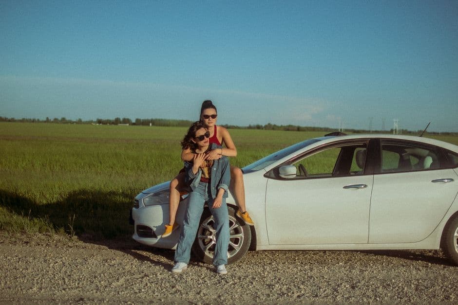 Two friends enjoying a sunny day by their car in an open field, showcasing friendship and travel vibes.